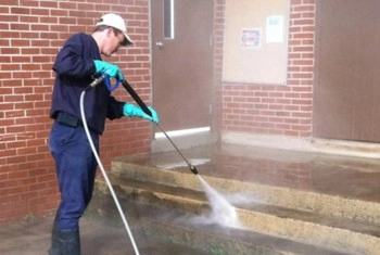 A person wearing gloves, boots, and a cap uses a pressure washer to clean concrete steps outside a brick building.
