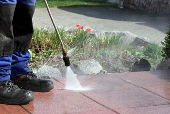 A person in blue pants and black shoes uses a pressure washer to clean red outdoor tiles, with garden plants and rocks visible in the background.