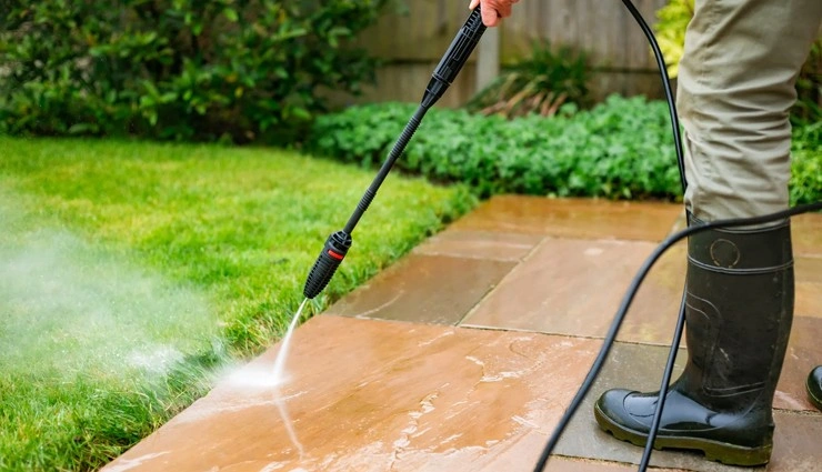 A person wearing boots uses a pressure washer to clean a stone patio next to a green lawn and garden. Water sprays forcefully from the nozzle, removing dirt from the patio surface.