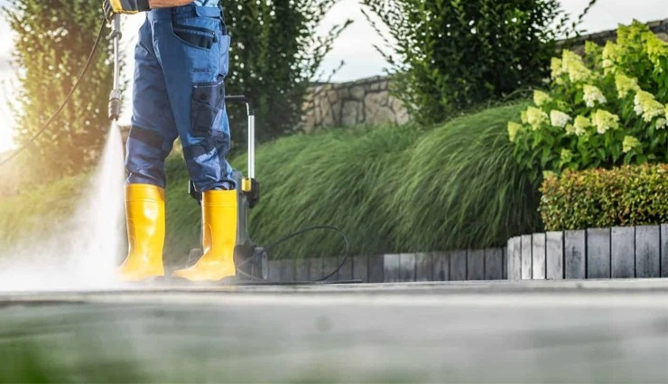 A person wearing blue pants and bright yellow rubber boots uses a power washer to clean an outdoor pavement, with green bushes and plants in the background.